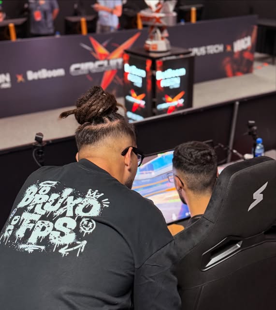 vinAo, de costas, com a camiseta O Bruxo do FPS, acompanhando um jogador numa bancada de torneio da Circuit X